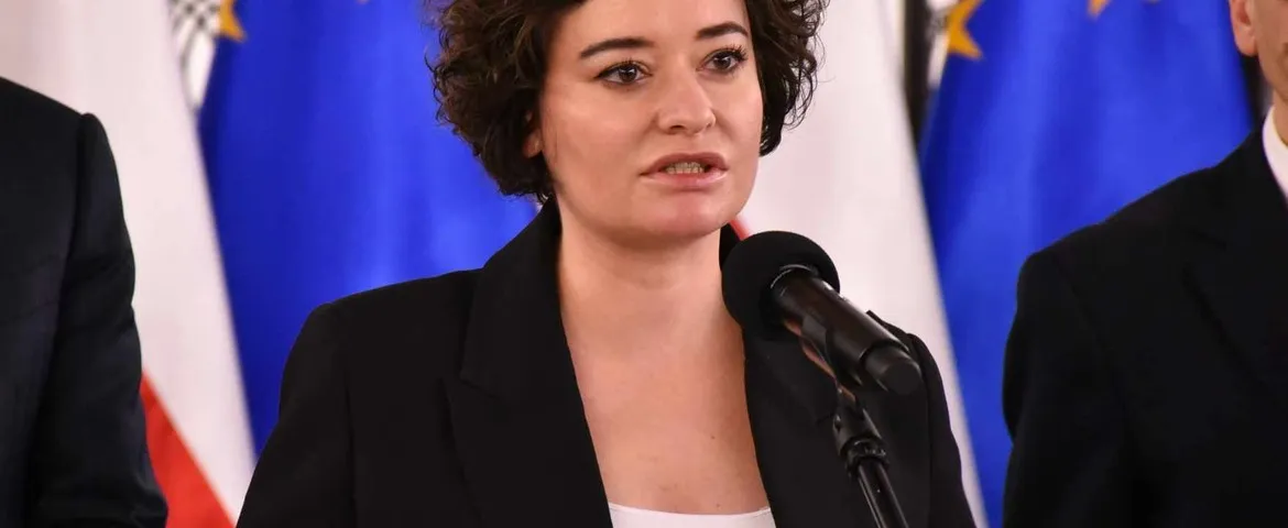 Kobieta w czarnym garniturze przemawia do mikrofonu podczas konferencji, w tle widoczne flagi.