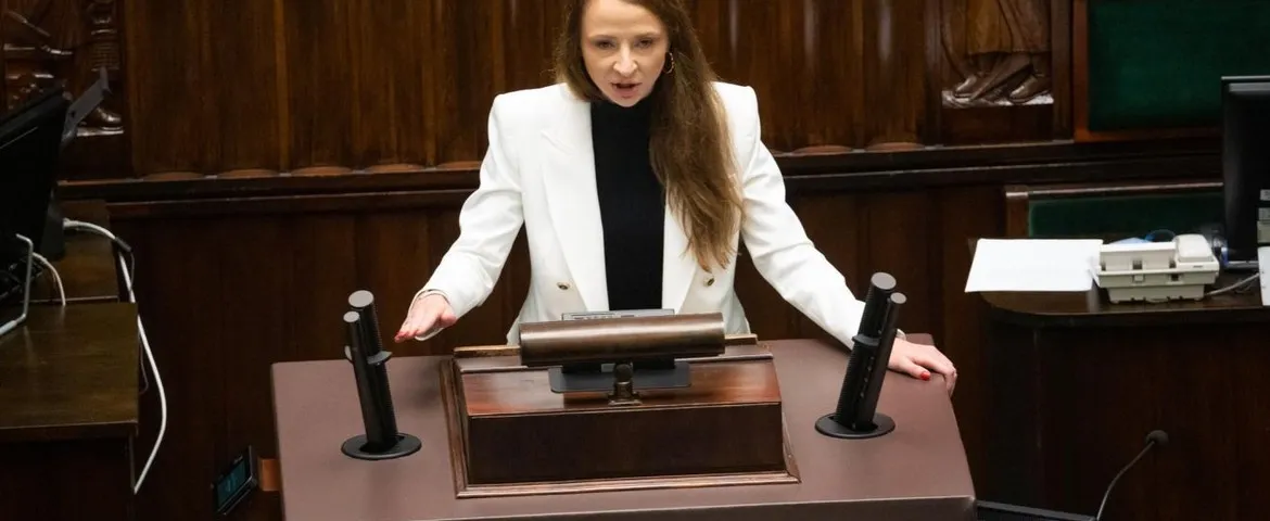 Kobieta w białym garniturze stoi za mównicą w sali obrad i wygłasza przemówienie do mikrofonów.