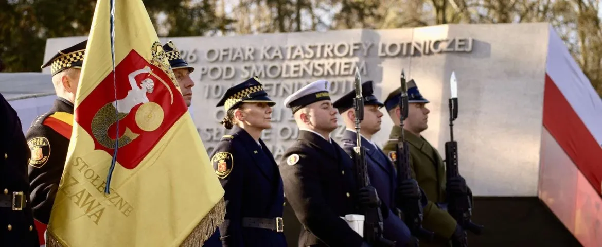 Grupa żołnierzy w mundurach stoi na ceremonii przed pomnikiem upamiętniającym ofiary katastrofy lotniczej pod Smoleńskiem, a na pierwszym planie widać flagę.