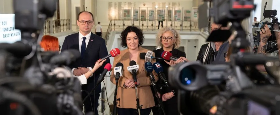 Grupa osób na konferencji prasowej w jasnym holu, pośrodku kobieta w beżowym płaszczu przemawia do zgromadzonych dziennikarzy otoczonych mikrofonami i kamerami.