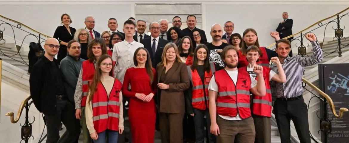 Duża grupa ludzi stojąca na schodach w eleganckim wnętrzu, część ubrana w czerwone kamizelki odblaskowe, wszyscy uśmiechnięci podczas wspólnego zdjęcia.