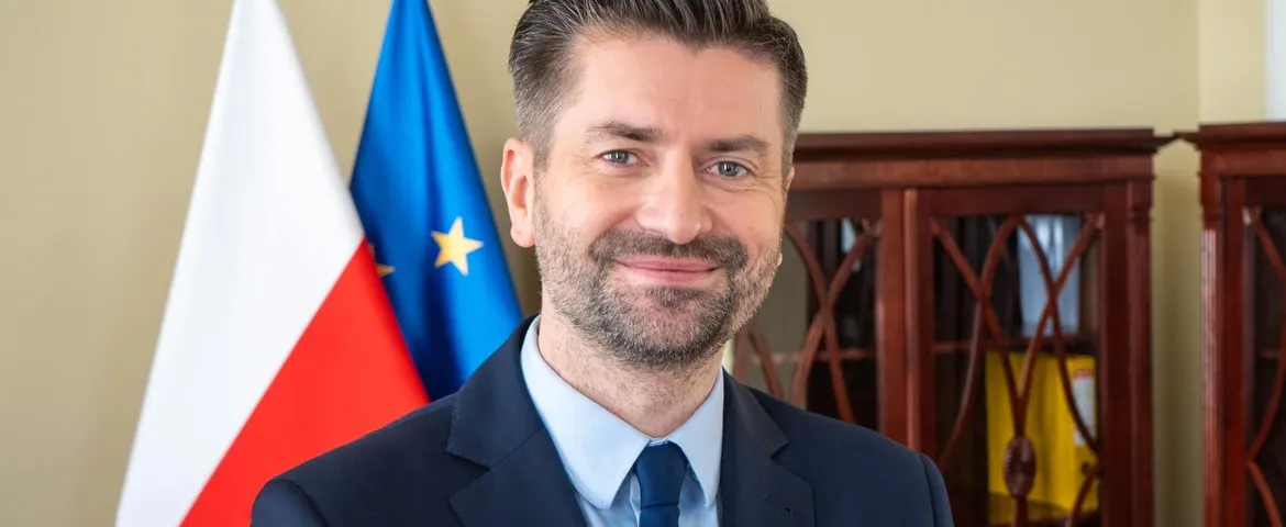 Mężczyzna w garniturze i niebieskim krawacie stoi na tle flag Polski i Unii Europejskiej, uśmiechnięty.