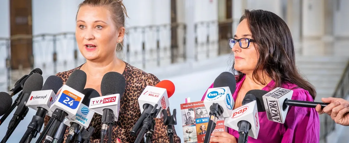 Dwie kobiety udzielają konferencji prasowej, stojąc przed licznymi mikrofonami dziennikarzy.