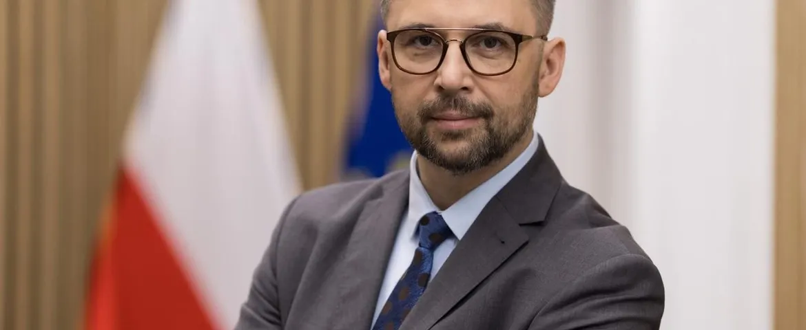 Mężczyzna w garniturze i okularach stoi w biurowym wnętrzu z flagami w tle.