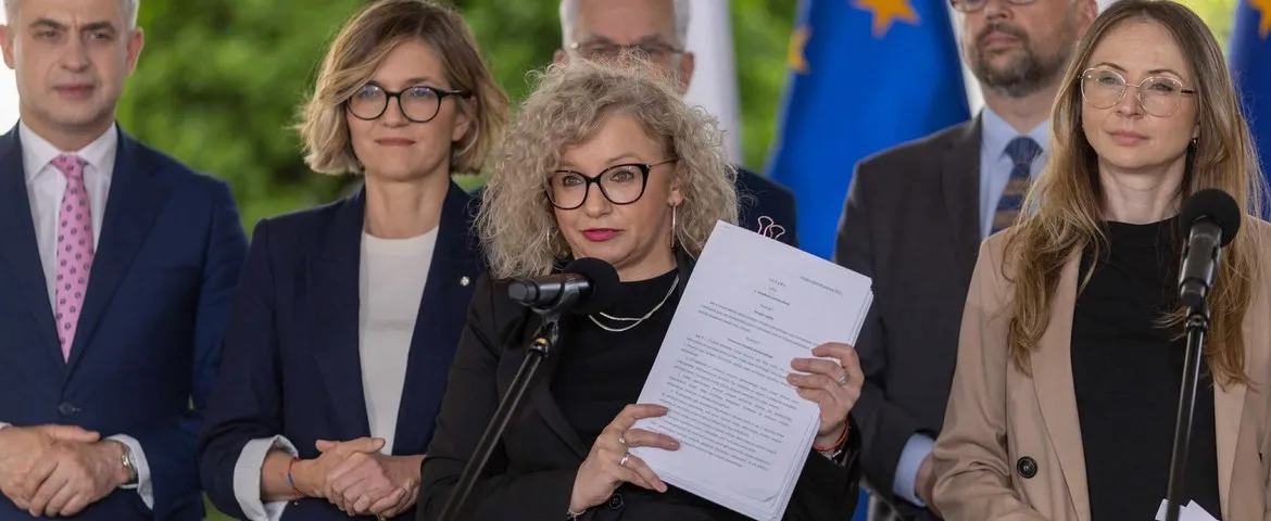 Grupa osób w formalnych strojach podczas konferencji prasowej; pośrodku kobieta z kręconymi blond włosami i okularami trzyma dokumenty przy mikrofonach, a w tle widać flagi.