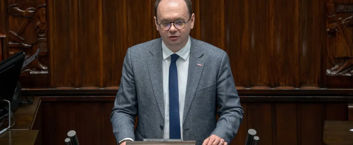 Mężczyzna w garniturze i okularach stoi przy podium w drewnianej sali posiedzeń i wygłasza przemówienie.