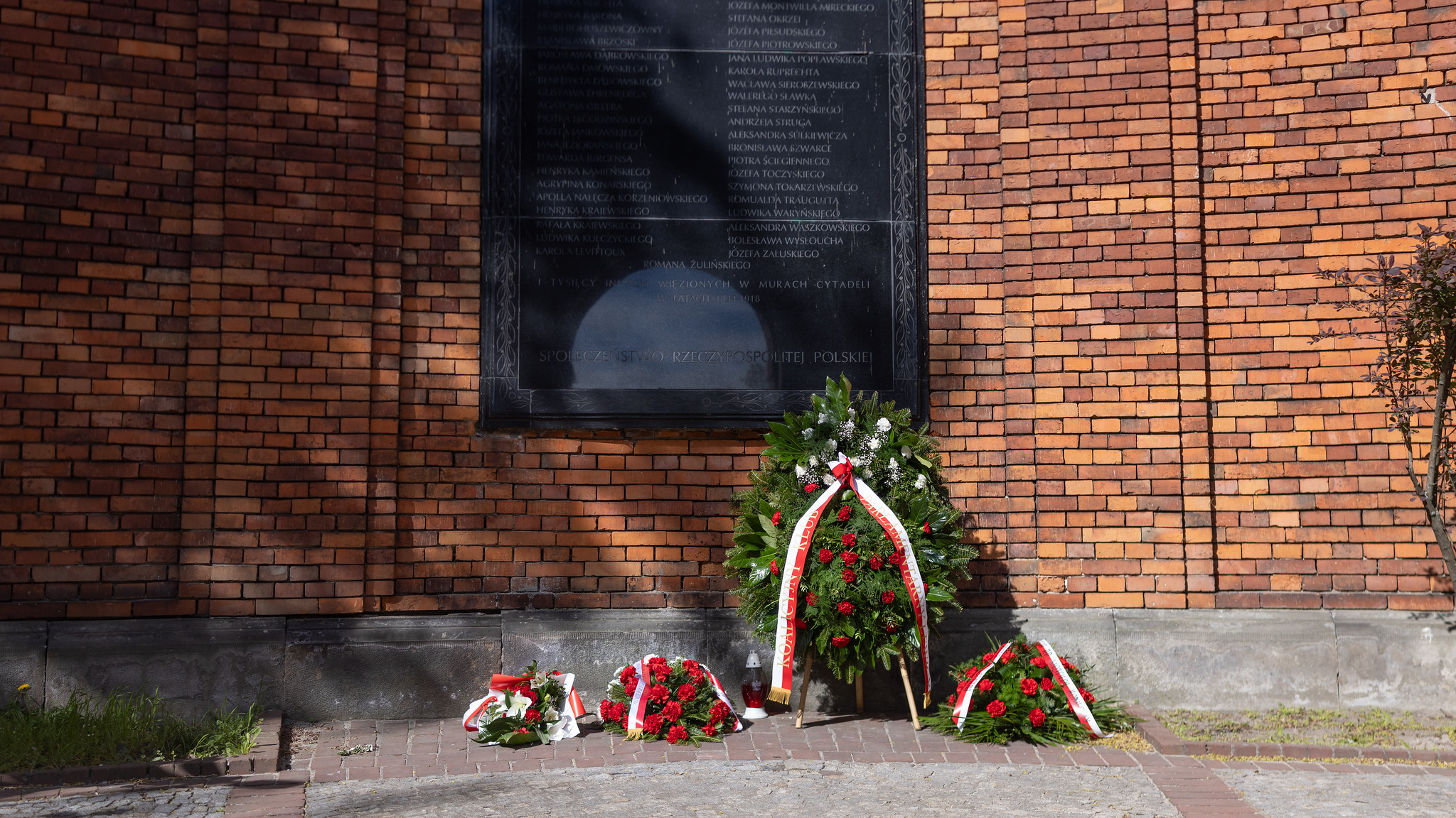Czarna tablica pamiątkowa zamontowana na ceglanej ścianie Cytadeli, z wieńcami kwiatów u podstawy.