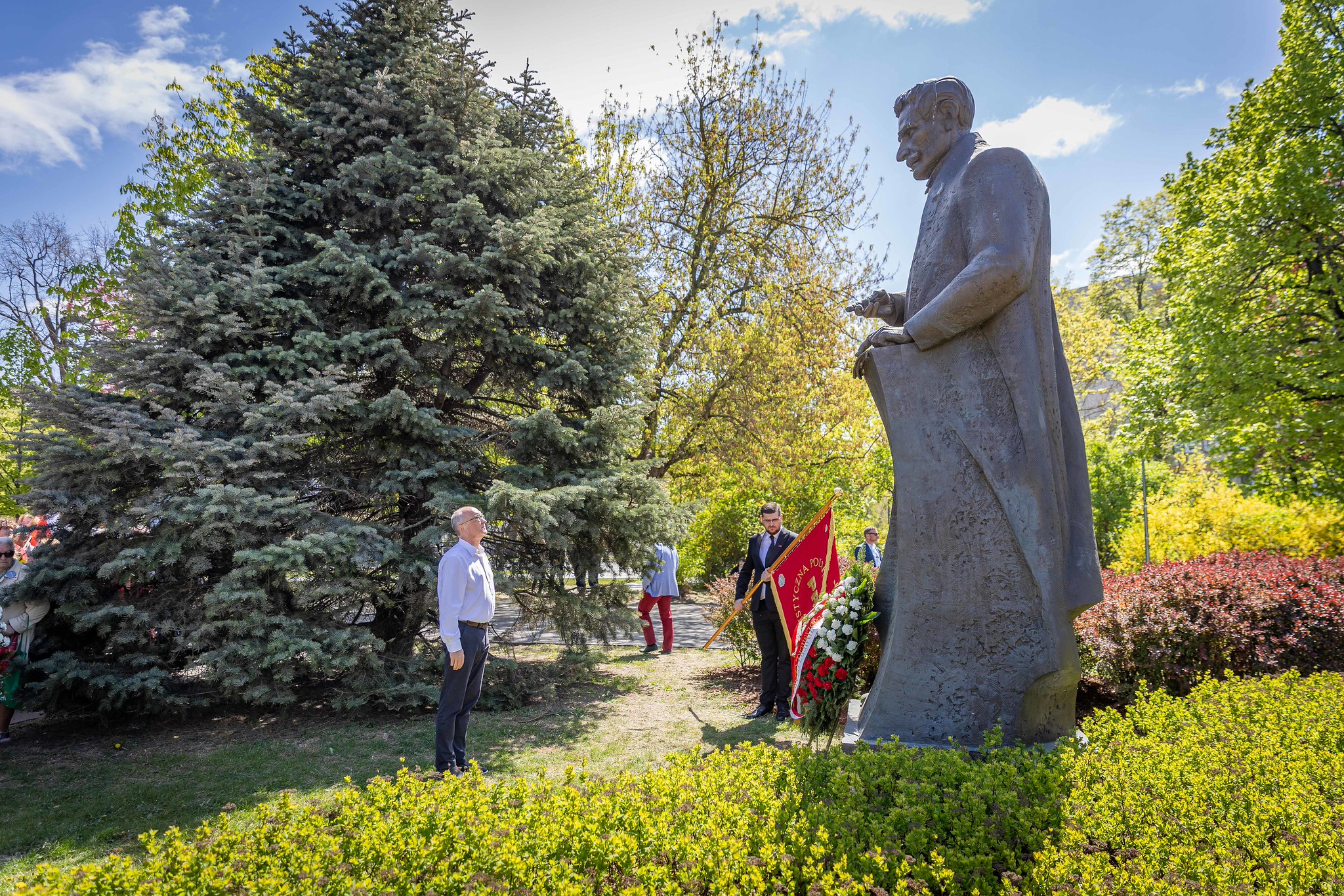 Posąg mężczyzny stoi w parku w otoczeniu zielonych drzew i krzewów, a kilku ludzi trzyma flagę i składa wieńce.