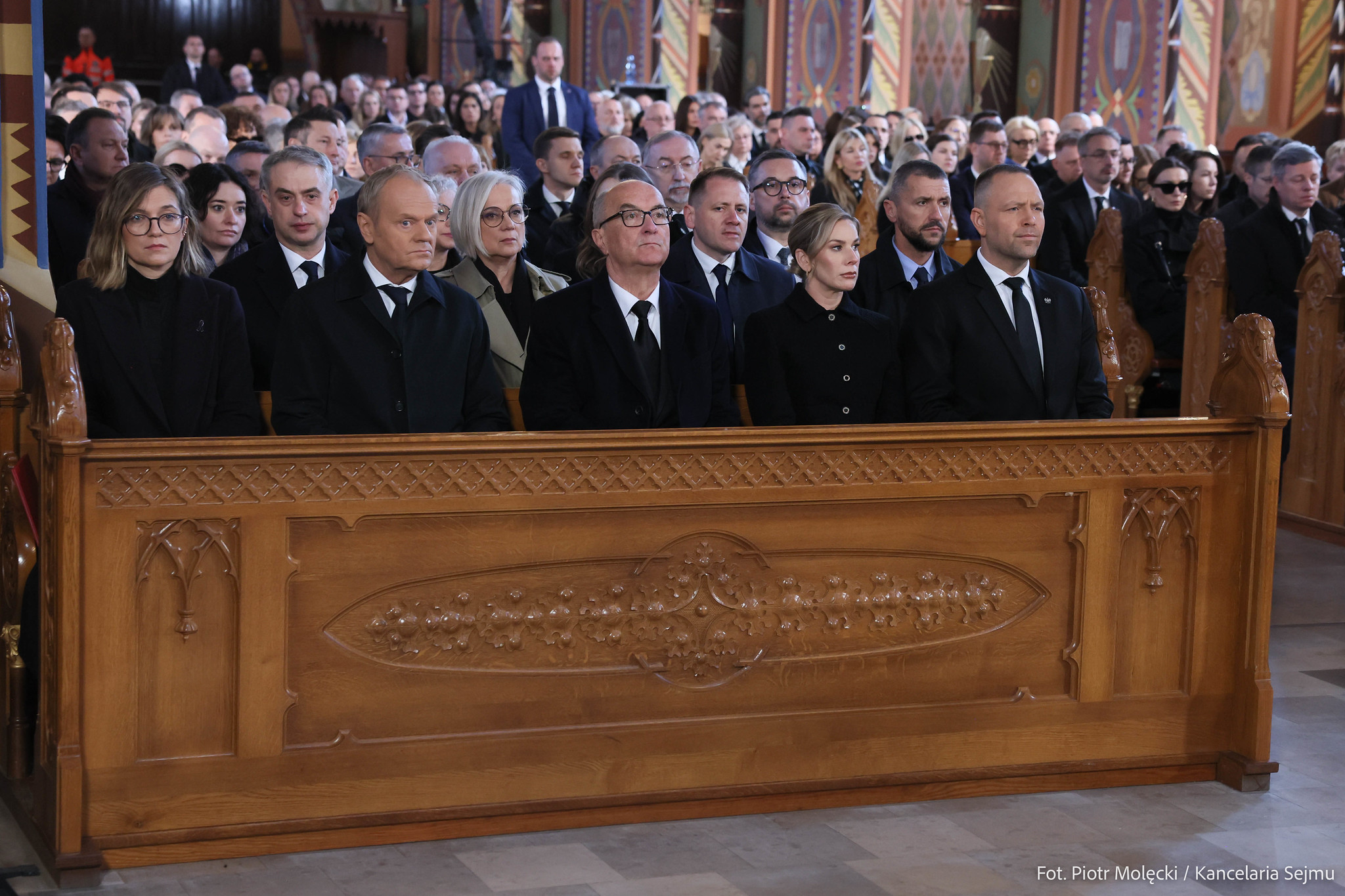 Grupa osób w formalnych ubraniach siedzi w ozdobnych drewnianych ławach w kościelnym wnętrzu podczas ceremonii.