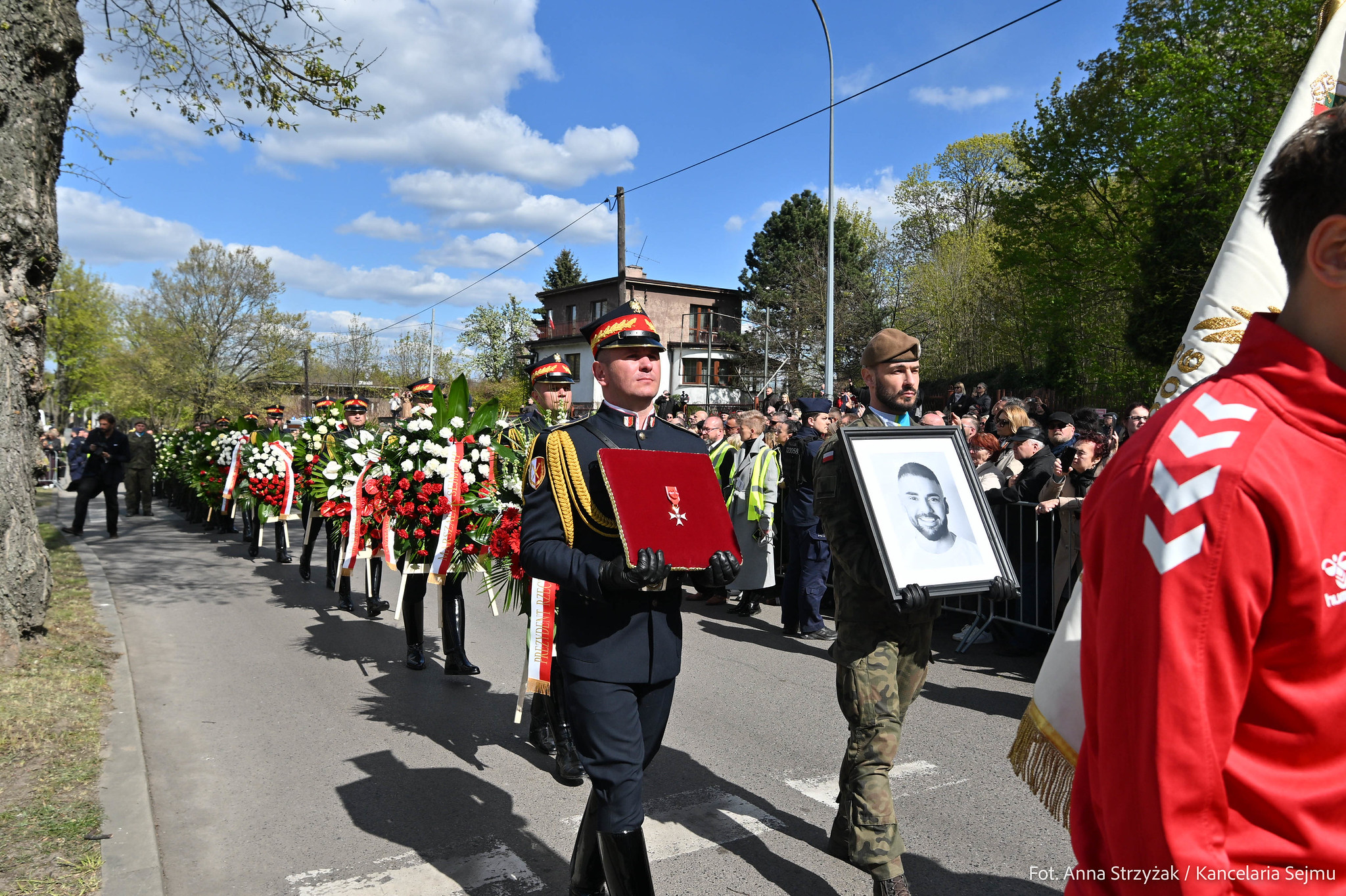 Żołnierze w eleganckich mundurach niosą kwietne wieńce i portret zmarłego podczas uroczystości upamiętniającej, otoczeni przez tłum i flagi na tle jasnego, pogodne nieba.