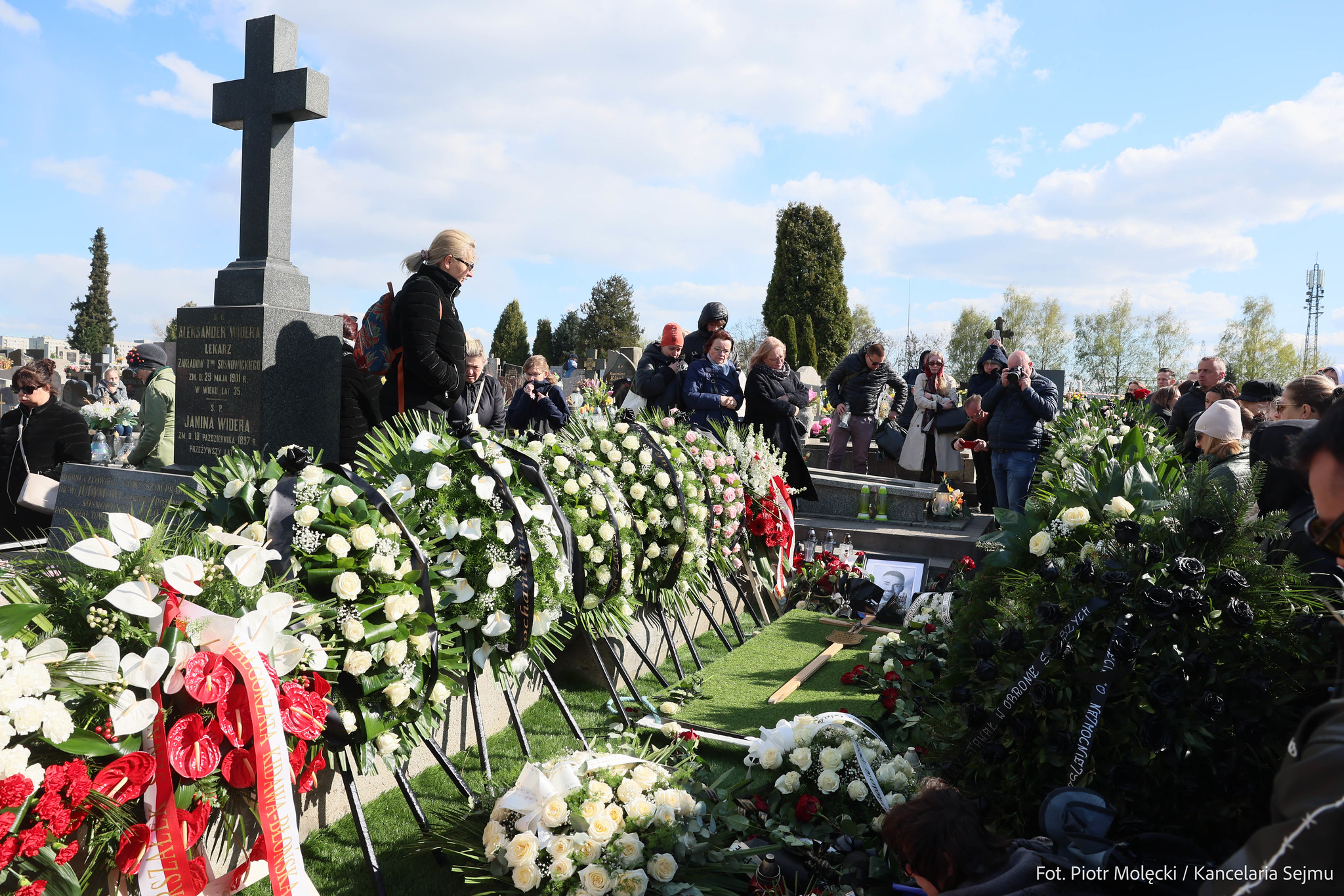 Grupa ludzi na cmentarzu składa kwiaty i modli się przy wysokim czarnym krzyżu, otoczonym nagrobkami i wieńcami.