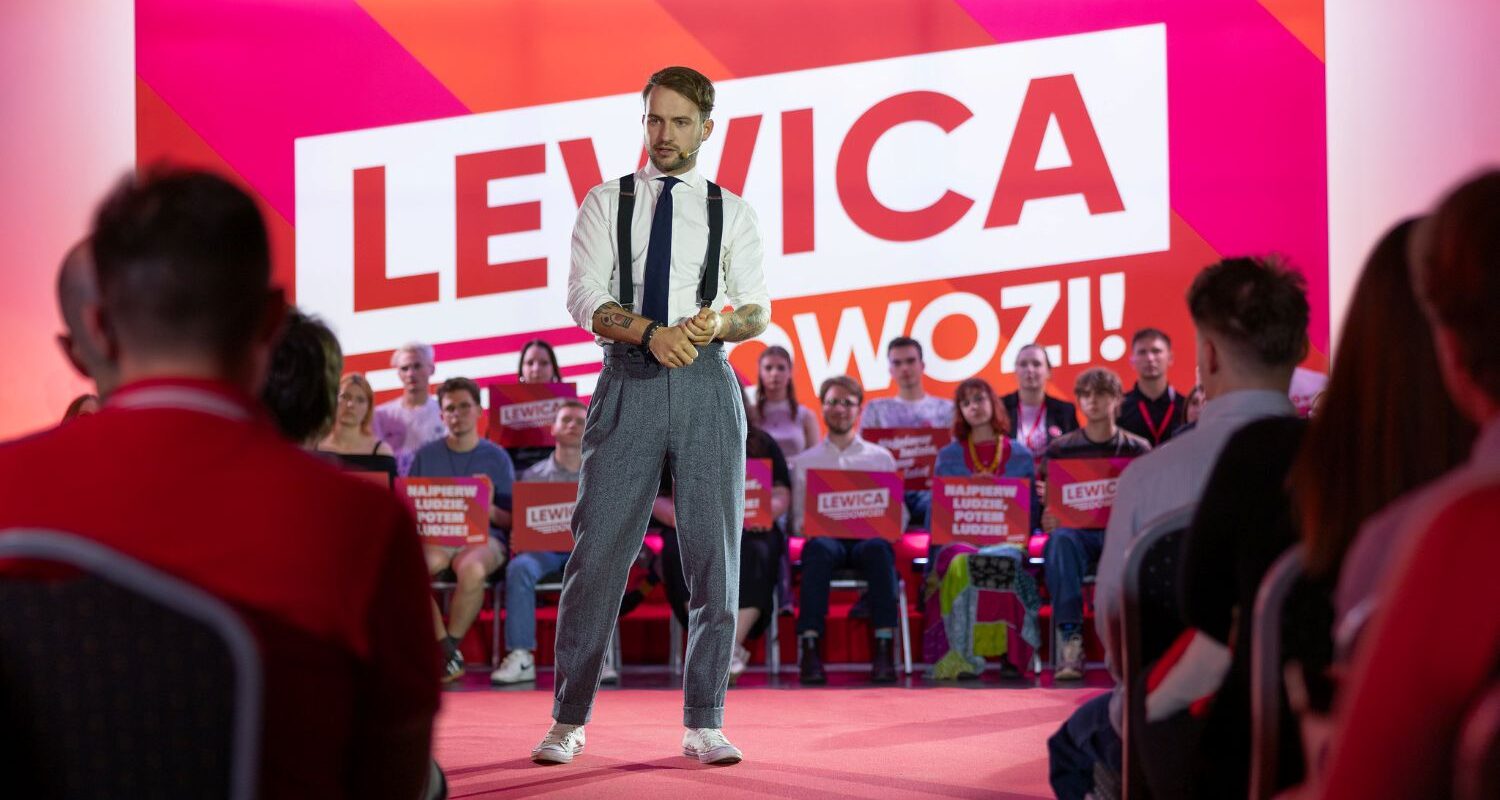 Mężczyzna w białej koszuli, czarnych suspendersach i szarych spodniach z tatuażami na ramionach stoi na scenie na czerwonym dywanie przed kolorową ścianą z napisem Lewica, otoczony publicznością trzymającą plakaty.