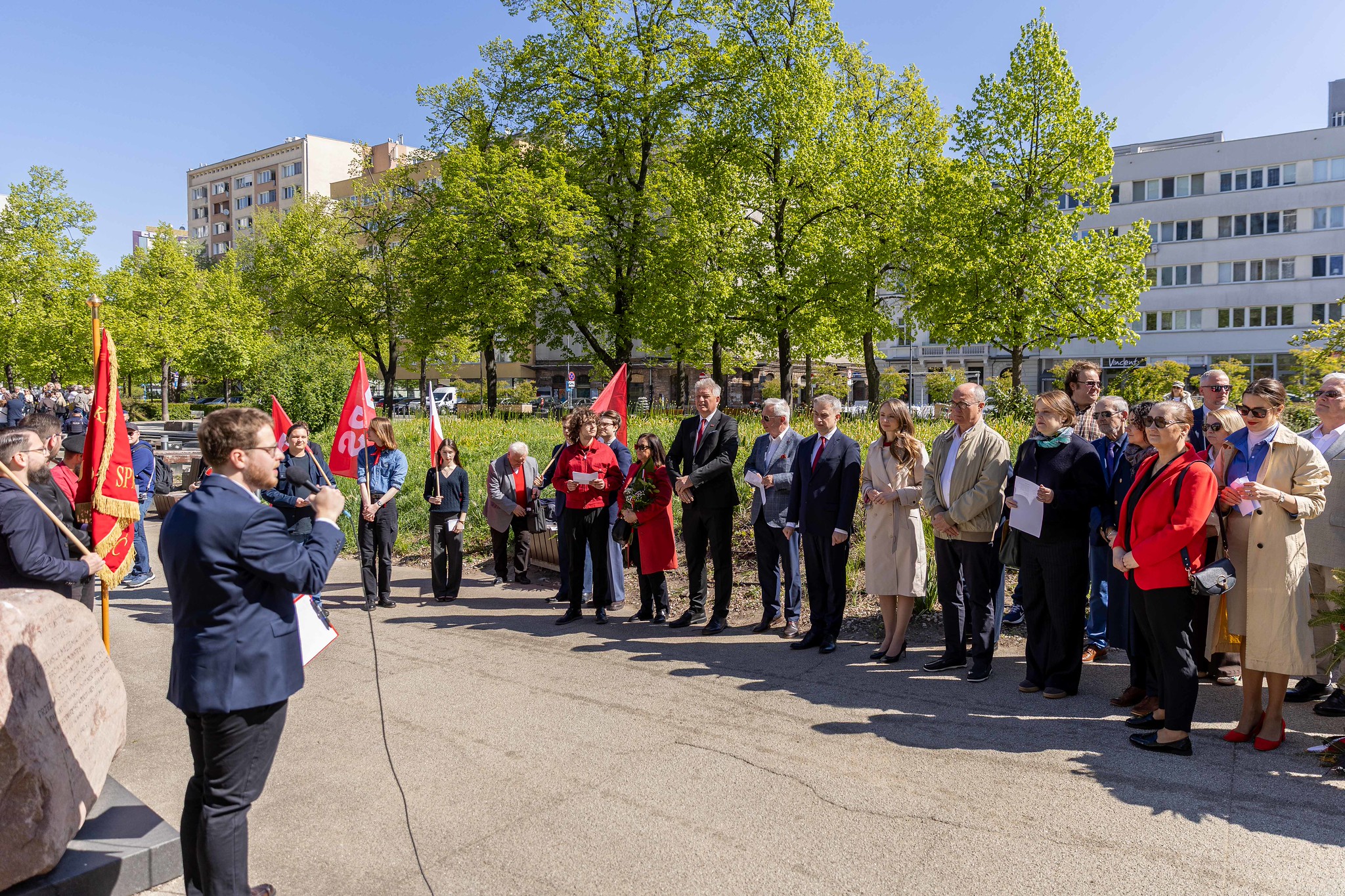 Grupa osób w eleganckich ubraniach stoi na placu podczas uroczystości, trzymając czerwone flagi, a w tle widać zielone drzewa i wielopiętrowe budynki.