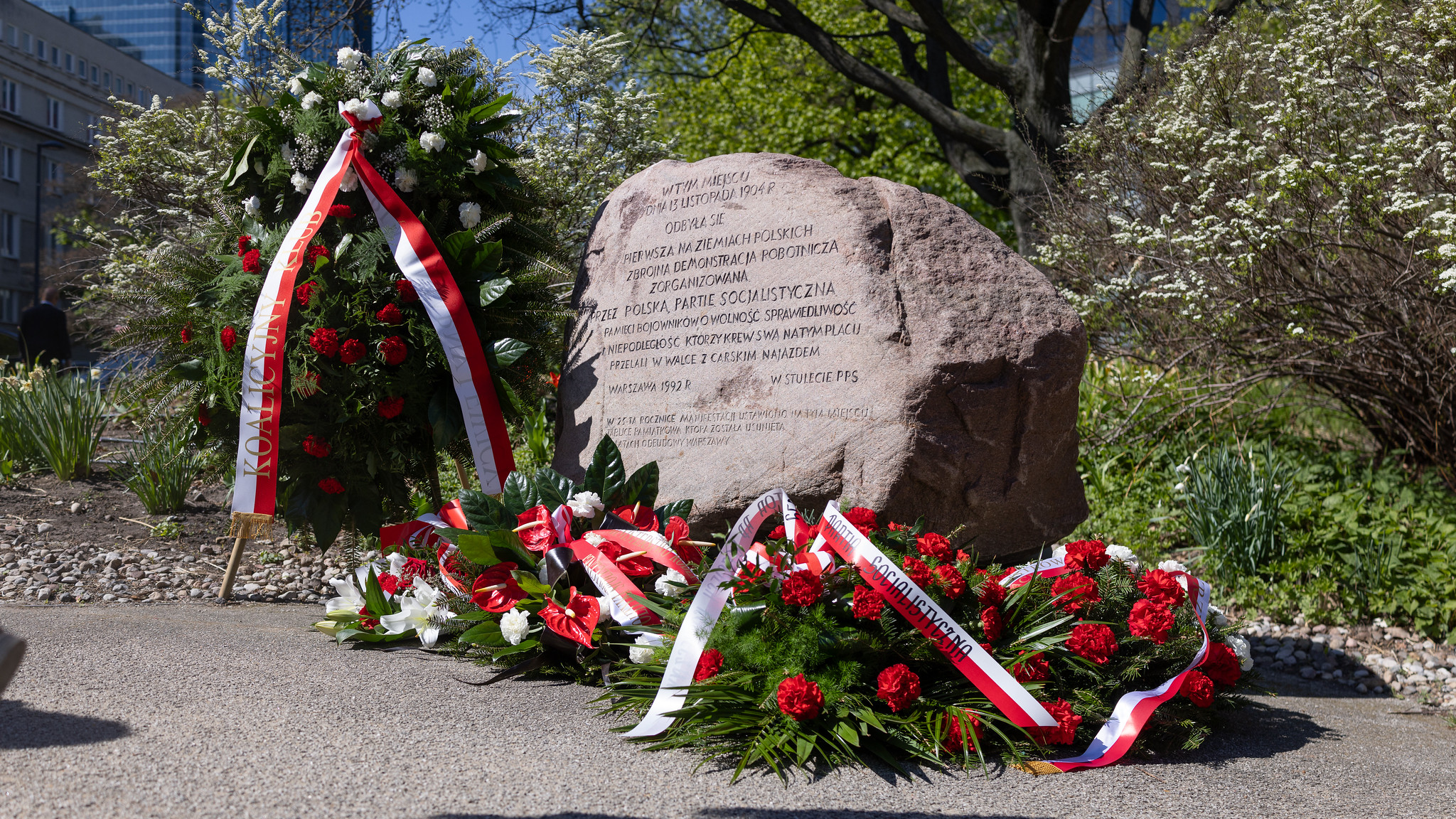 Kamienny obelisk z wyrytym tekstem, otoczony wieńcami z czerwonych i białych kwiatów oraz czerwono-białymi wstążkami, na Placu Grzybowskich.