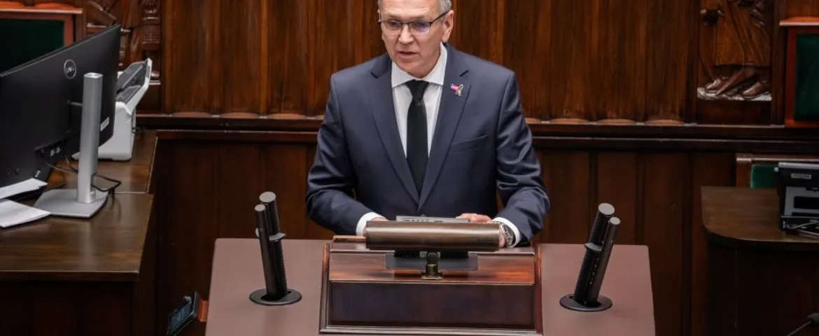 Mężczyzna w garniturze stoi za mównicą w drewnianej sali, między dwoma mikrofonami, wygłaszając przemówienie.