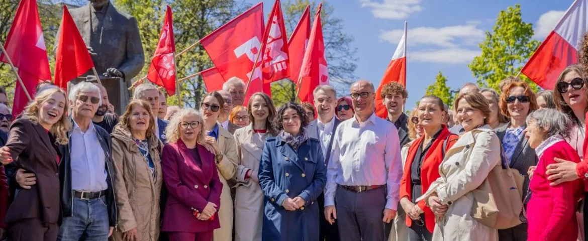 Grupa ludzi stojąca na zewnątrz podczas wydarzenia, trzymających czerwono-białe flagi, w tle widoczna statua i zielone drzewa pod błękitnym niebem.