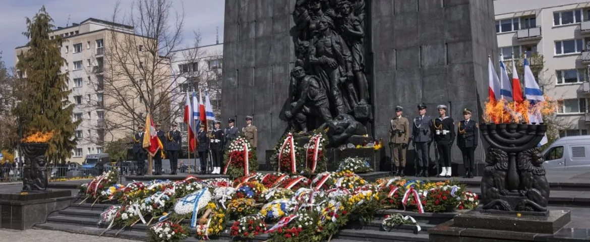 Uroczystość przy pomniku z rzeźbami, żołnierze w mundurach stoją z flagami, wokół kwietne wieńce, a po obu stronach płoną ceremonialne ogniska.