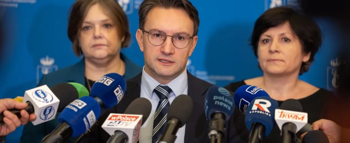 Mężczyzna w garniturze i okularach przemawia do licznych mikrofonów podczas konferencji prasowej, stojąc przed niebieskim tłem, za którym stoją dwie kobiety.