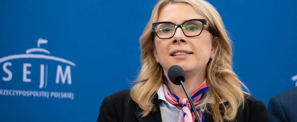 Kobieta z blond włosami i okularami przemawia do mikrofonu na tle niebieskiej ściany z logo Sejmu.