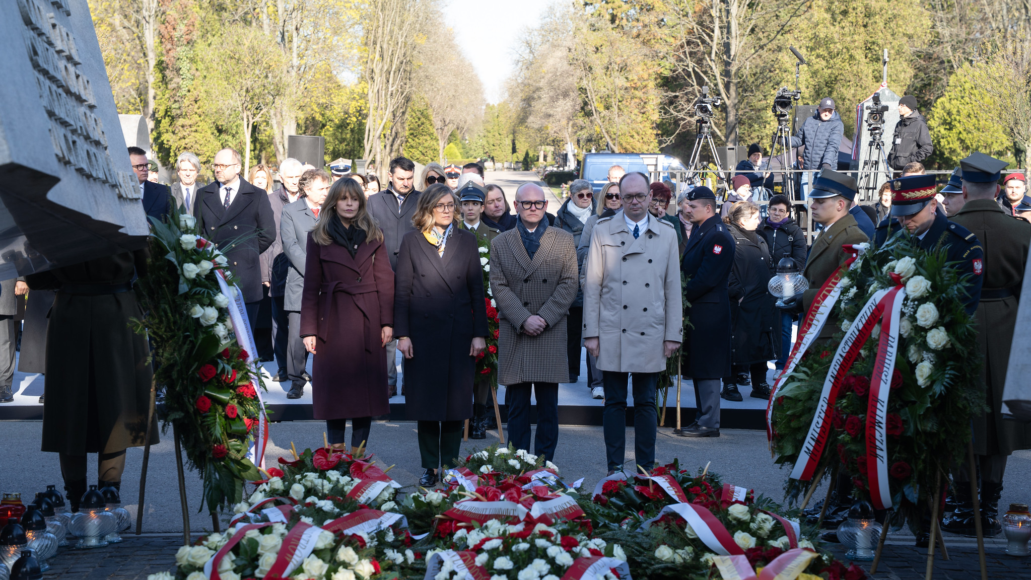 Grupa osób w eleganckich ubraniach i mundurach bierze udział w uroczystości upamiętniającej katastrofę smoleńską, otoczona kwiatami i kamerami.