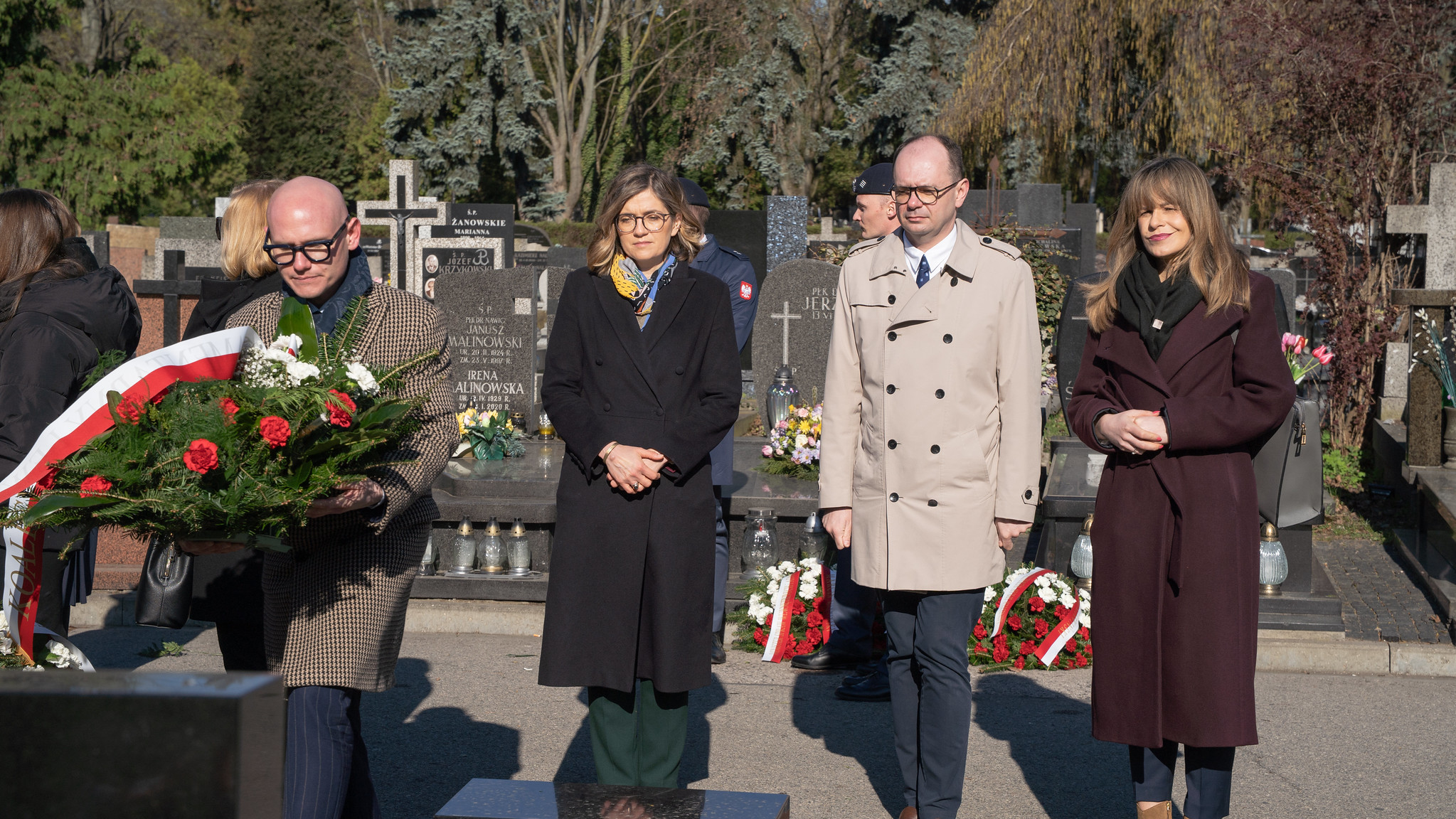 Grupa osób w eleganckich ubraniach stoi na cmentarzu obok grobów, trzyma kwiaty podczas ceremonii upamiętniającej.