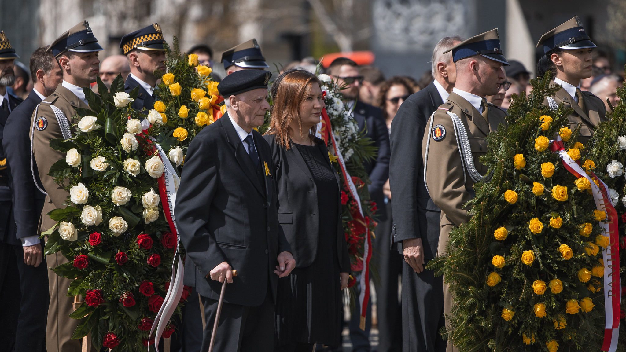 Grupa osób w mundurach wojskowych i w cywilnych ubraniach stoi podczas ceremonii, trzymając duże wieńce z żółtych i białych róż oraz gałęzi zieleni.