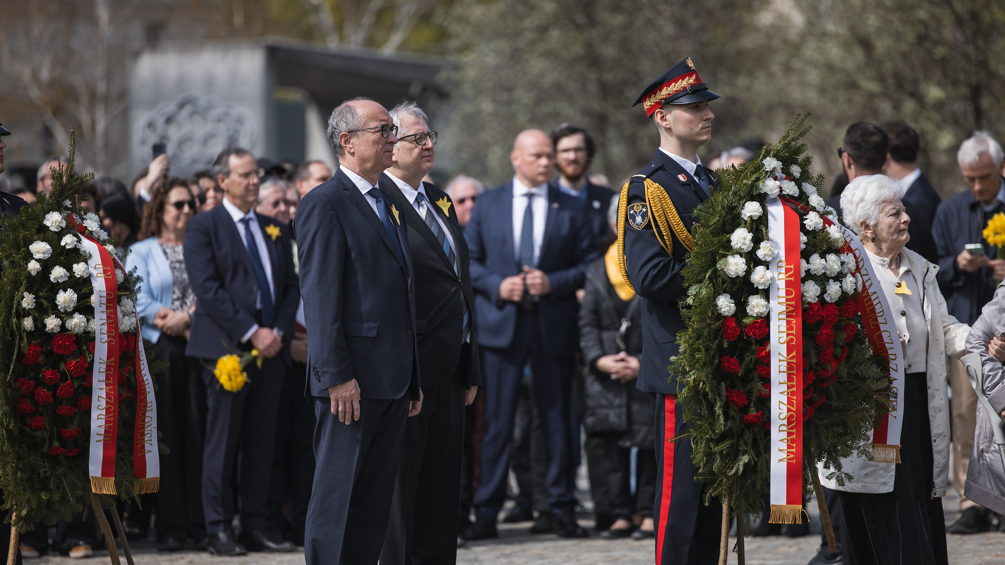 Grupa uczestników ceremonii stoi w garniturach i mundurach, trzymając duże wieńce z białych i czerwonych kwiatów ozdobione wstęgami.