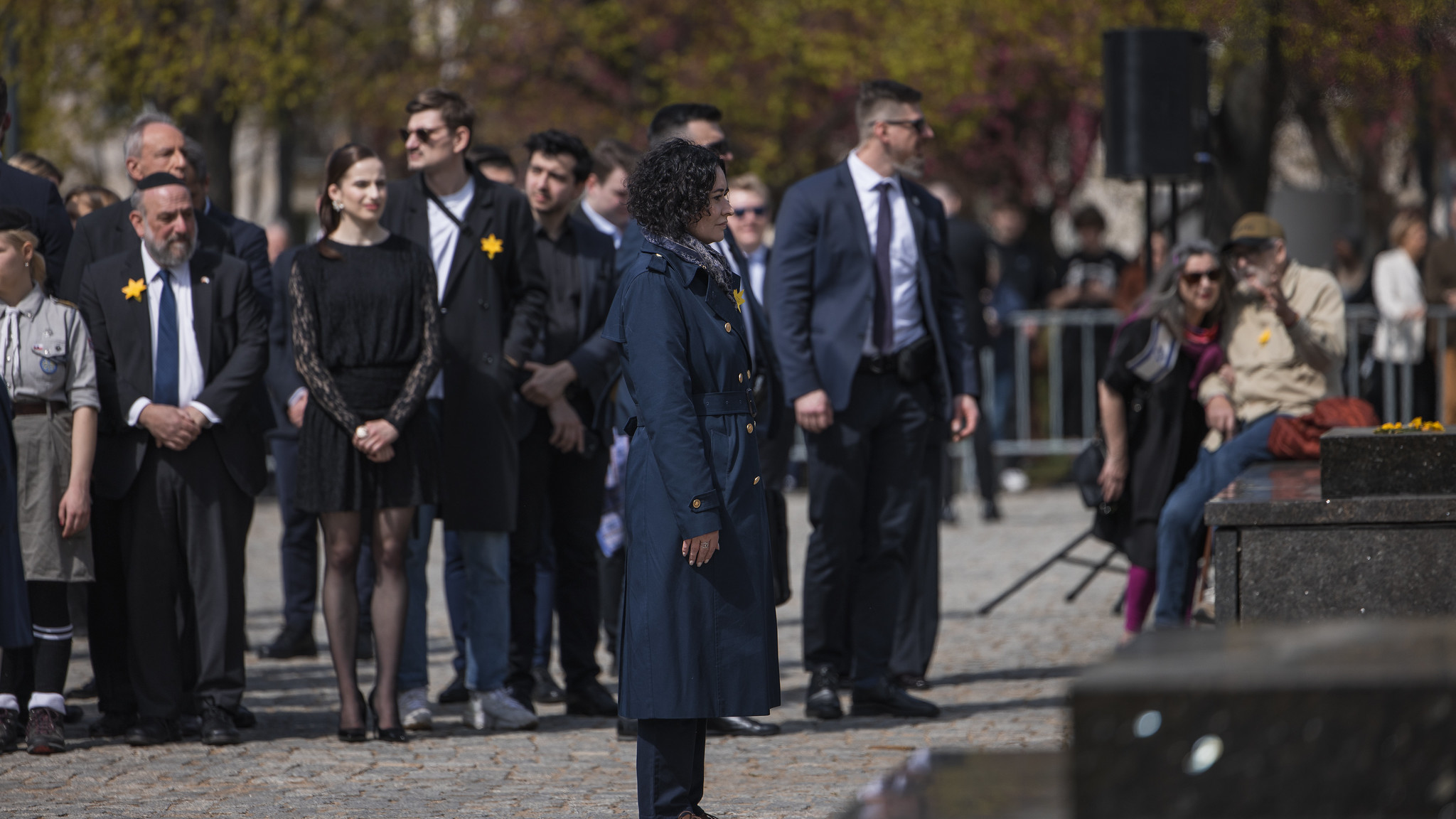 Kobieta w granatowym płaszczu stoi na pierwszym planie wśród grupy ludzi ubranych w eleganckie stroje podczas wydarzenia na placu, wszyscy mają żółte kwiaty przypięte do ubrań.